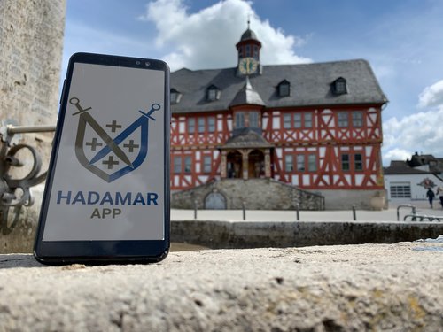 Ein Smartphone mit einem Hadamar-App-Logo vor dem Hadamarer Rathaus